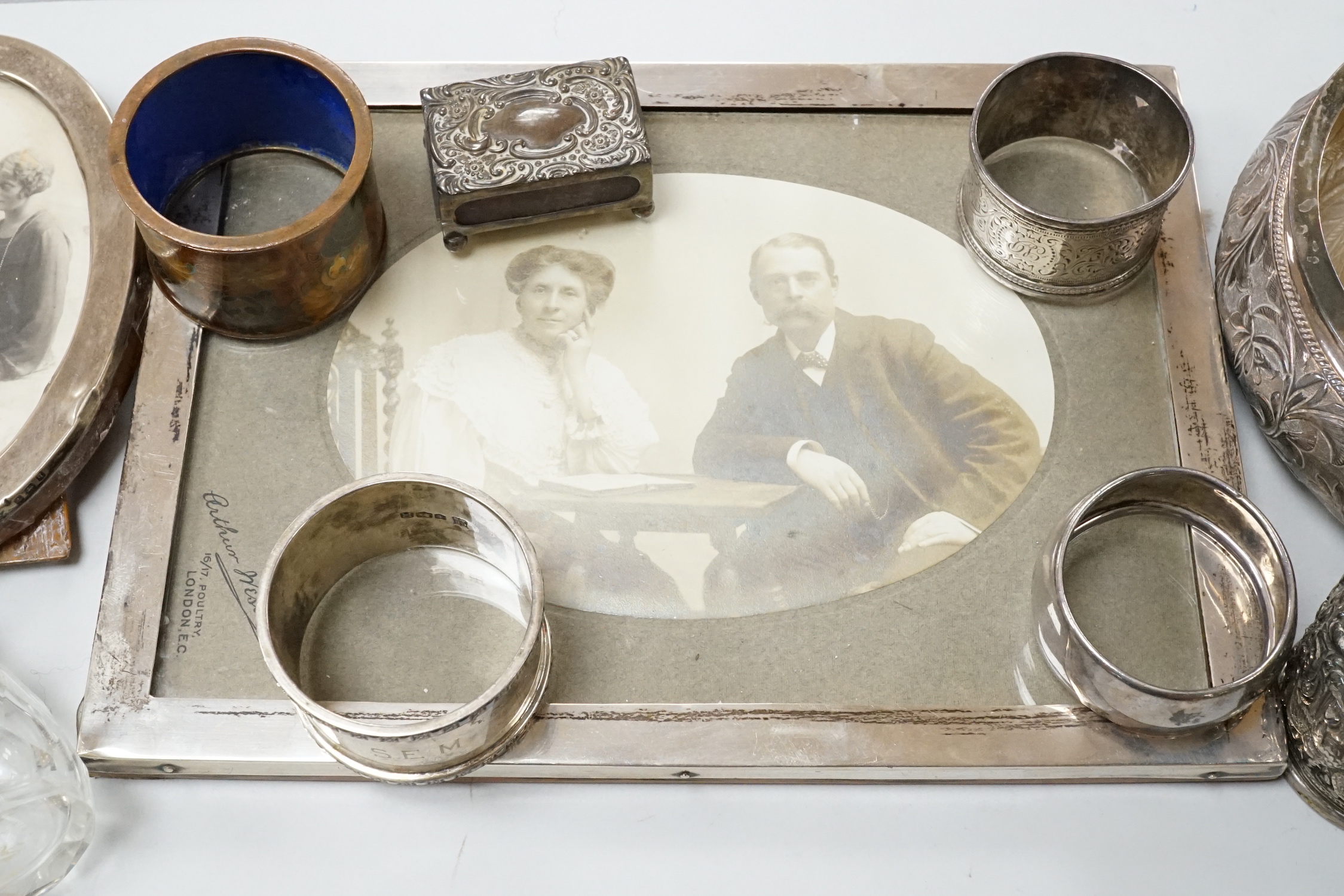 Two George V silver mounted photograph frames including oval, an Indian white metal flower holder and other items including silver napkin rings and a matchbox sleeve.